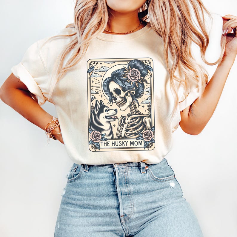 May include: Cream-colored t-shirt featuring a tarot card-style graphic. The graphic depicts a skeleton with a floral hairpiece and a husky dog. The text "THE HUSKY MOM" is at the bottom of the graphic. The shirt is paired with blue jeans.