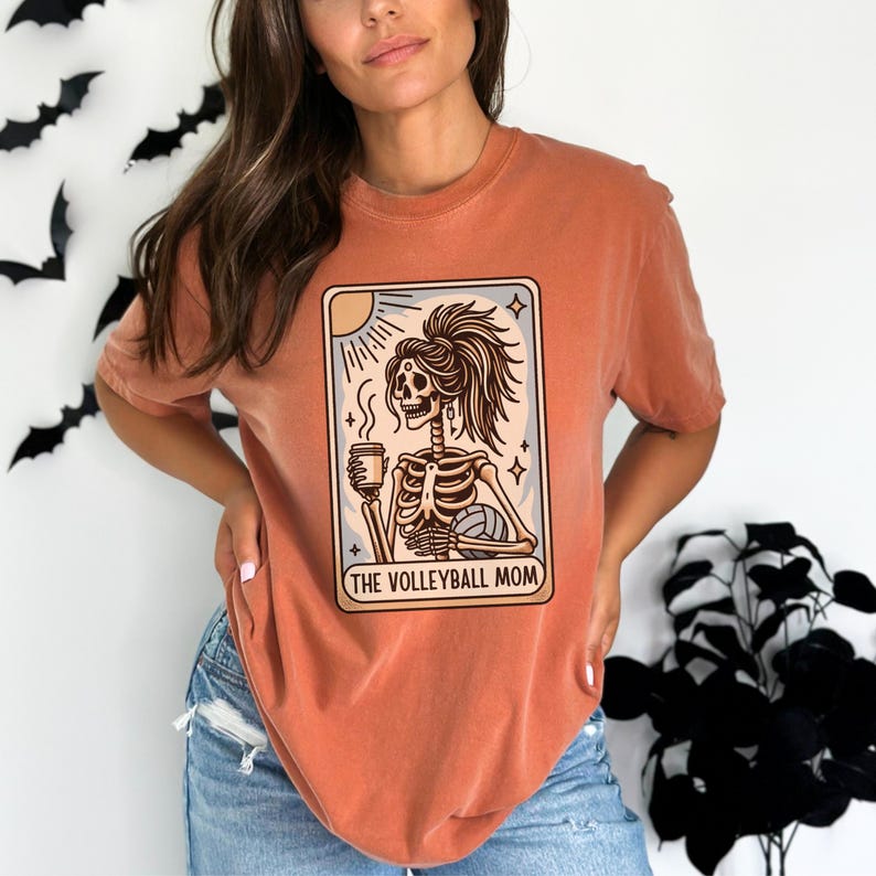 Może przedstawiać: Pomarańczowy T-shirt z grafiką przedstawiającą szkielet trzymający kubek kawy i piłkę do siatk&oacute;wki. Grafika jest w stylu karty tarota z tekstem "THE VOLLEYBALL MOM".
