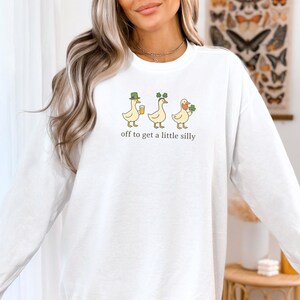 May include: White sweatshirt featuring a whimsical graphic of three ducks, each adorned with St. Patrick's Day themed accessories. The ducks are holding a beer, shamrocks, and a shamrock. The text "off to get a little silly" is printed below the ducks.