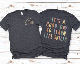 May include: A dark heather grey t-shirt with a sun graphic and the text "FCS Teacher" on the front. The back of the shirt has the text "It's a good day to learn life skills" in colorful letters.
