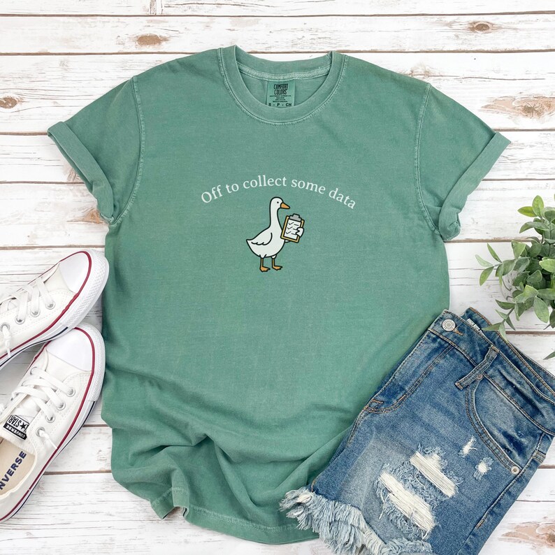 May include: A sage green t-shirt with rolled-up sleeves featuring the phrase "Off to collect some data" and a cartoon goose holding a clipboard. White sneakers and denim shorts are also visible.