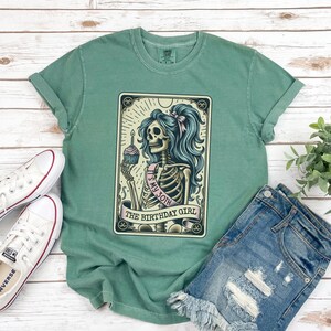 May include: A green t-shirt featuring a vintage-style illustration of a skeleton holding a cupcake with a lit candle. The skeleton is wearing a sash that says "It's My 30th" and the text "The Birthday Girl" is below the image.