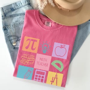 May include: A pink t-shirt with a math teacher theme. The shirt features colorful squares with math symbols, an apple, a lightbulb, books, a pencil, a calculator, and a protractor. The text "MATH TEACHER" is also printed on the shirt.