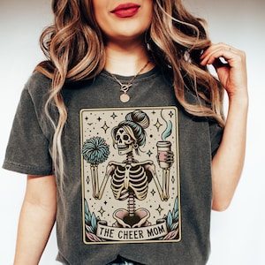 Pode incluir: Uma camiseta cinza escura com uma ilustra&ccedil;&atilde;o vintage de um esqueleto segurando uma x&iacute;cara de caf&eacute; e um pompom. A ilustra&ccedil;&atilde;o &eacute; cercada por uma borda bege e o texto "THE CHEER MOM" &eacute; impresso abaixo da imagem.