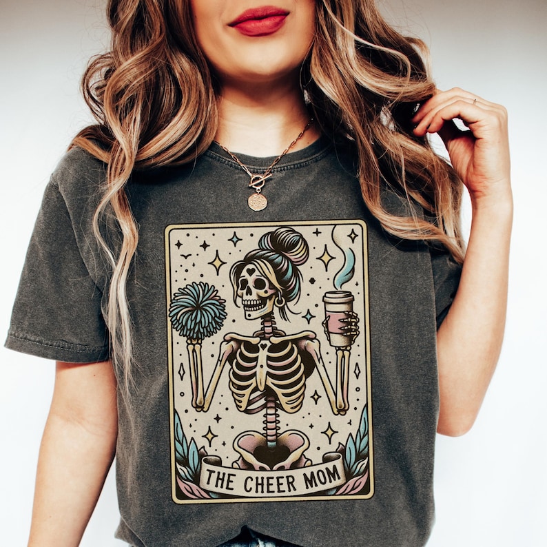 Pode incluir: Uma camiseta cinza escura com uma ilustra&ccedil;&atilde;o vintage de um esqueleto segurando uma x&iacute;cara de caf&eacute; e um pompom. A ilustra&ccedil;&atilde;o &eacute; cercada por uma borda bege e o texto "THE CHEER MOM" &eacute; impresso abaixo da imagem.