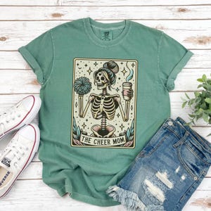 Pode incluir: Camiseta verde com uma ilustra&ccedil;&atilde;o vintage de um esqueleto segurando uma x&iacute;cara de caf&eacute; e um pompom de l&iacute;der de torcida. O texto "The Cheer Mom" est&aacute; impresso abaixo da ilustra&ccedil;&atilde;o.