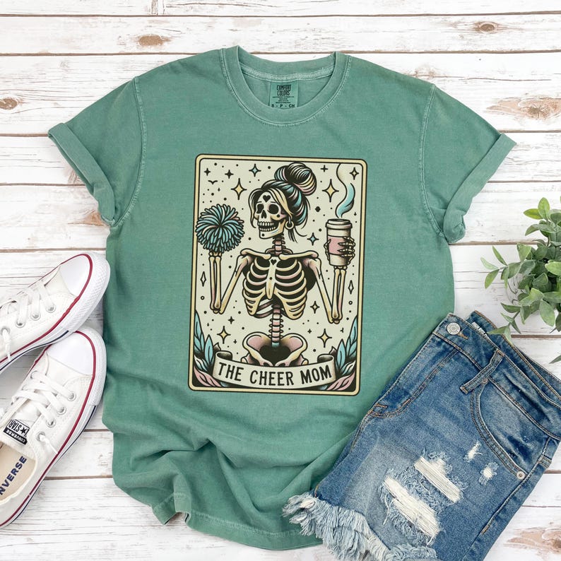 Pode incluir: Camiseta verde com uma ilustra&ccedil;&atilde;o vintage de um esqueleto segurando uma x&iacute;cara de caf&eacute; e um pompom de l&iacute;der de torcida. O texto "The Cheer Mom" est&aacute; impresso abaixo da ilustra&ccedil;&atilde;o.