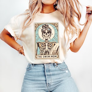 May include: A white t-shirt with a graphic of a skeleton wearing sunglasses and holding a surfboard. The graphic is in a vintage tarot card style and says "THE SWIM MOM".