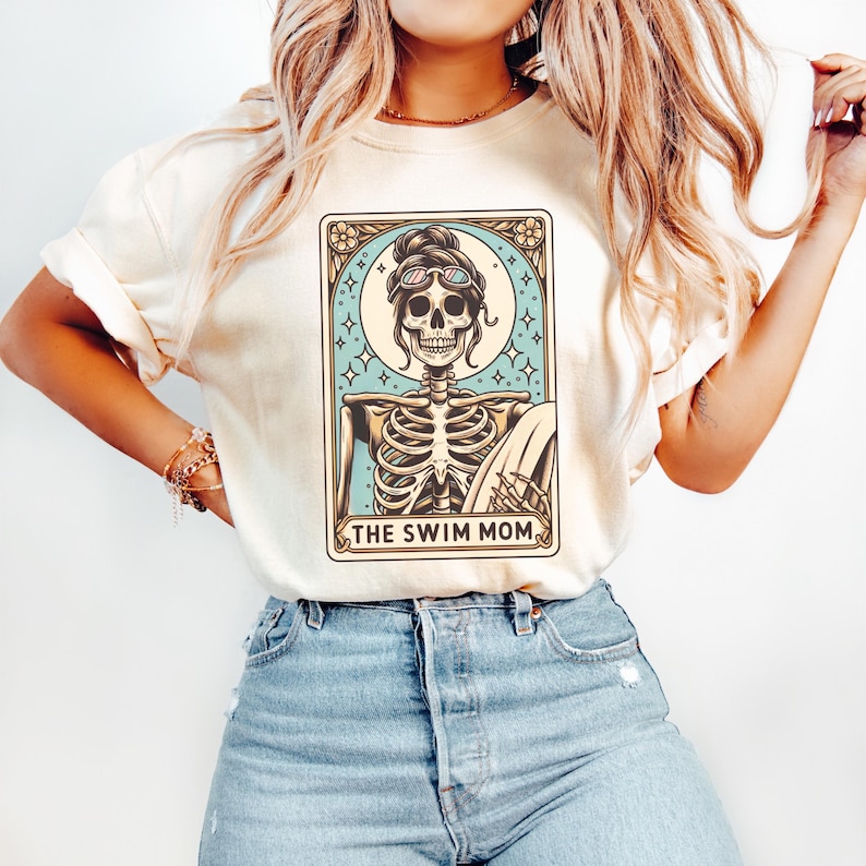 May include: A white t-shirt with a graphic of a skeleton wearing sunglasses and holding a surfboard. The graphic is in a vintage tarot card style and says "THE SWIM MOM".