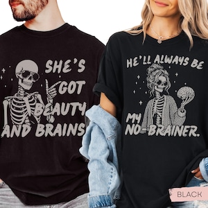 Może przedstawiać: Dwie czarne koszulki z grafikami. Lewa koszulka przedstawia szkielet w okularach przeciwsłonecznych z napisem SHE'S GOT BEAUTY AND BRAINS. Prawa koszulka pokazuje szkielet w koku i kardiganie trzymający mózg z napisem HE'LL ALWAYS BE MY NO-BRAINER.