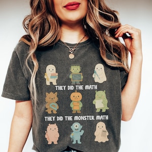May include: Dark gray t-shirt featuring cartoon monsters with math-related props. The text "THEY DID THE MATH" and "THEY DID THE MONSTER MATH" are printed below the monsters. The shirt has a crew neck and short sleeves.