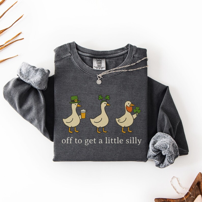 May include: Dark gray sweatshirt with a whimsical design of three ducks, each adorned with St. Patrick's Day themed accessories. The ducks are holding a beer, shamrocks, and a leprechaun beard. The text "off to get a little silly" is printed below the ducks.
