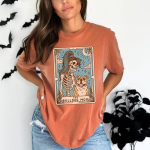 May include: A burnt orange t-shirt featuring a graphic of a skeleton woman with long black hair holding a bulldog. The graphic is in a tarot card style with the text "The Bulldog Mom" at the bottom.