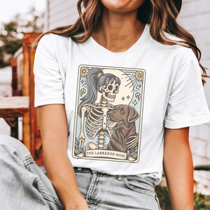 May include: White t-shirt with a graphic of a skeleton woman holding a brown labrador retriever. The graphic is in a vintage style with a light blue background and the text "The Labrador Mom".