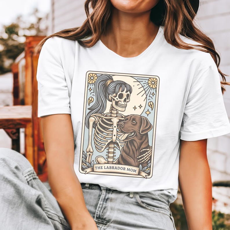 May include: White t-shirt with a graphic of a skeleton woman holding a brown labrador retriever. The graphic is in a vintage style with a light blue background and the text "The Labrador Mom".