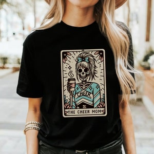 Pu&ograve; includere: Maglietta nera con una grafica di uno scheletro che indossa un costume da cheerleader e tiene una tazza di caff&egrave;. La grafica &egrave; in stile vintage e include il testo "The Cheer Mom".
