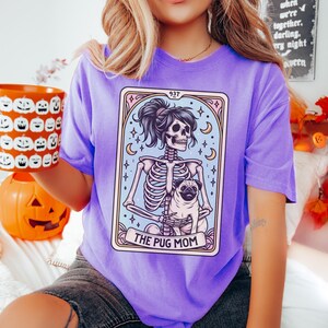 Puede incluir: Camiseta morada con un gr&aacute;fico de un esqueleto sosteniendo un pug. El gr&aacute;fico est&aacute; en el estilo de una carta de tarot y dice "The Pug Mom" en la parte inferior.