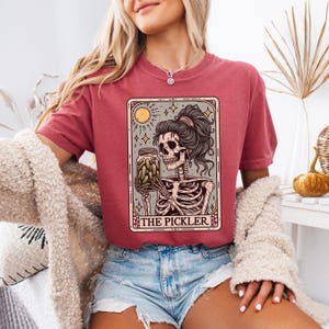 Pode incluir: Uma camiseta vermelha com um gr&aacute;fico de um esqueleto segurando um pote de picles. O gr&aacute;fico est&aacute; em estilo de carta de tar&ocirc; com o texto "The Pickler" na parte inferior.