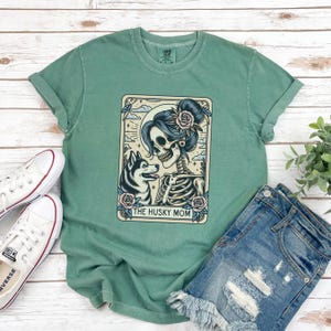 May include: A seafoam green t-shirt featuring a graphic of a skeleton with a husky dog and the text "THE HUSKY MOM". The graphic is in a rectangular frame with floral and celestial elements. The shirt is paired with denim shorts and white sneakers.