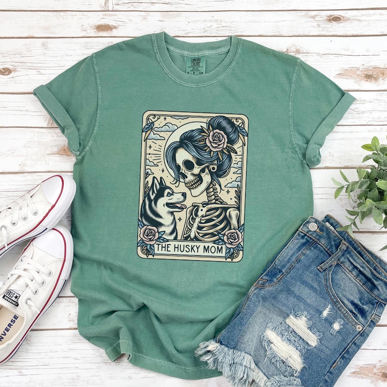 May include: A seafoam green t-shirt featuring a graphic of a skeleton with a husky dog and the text "THE HUSKY MOM". The graphic is in a rectangular frame with floral and celestial elements. The shirt is paired with denim shorts and white sneakers.