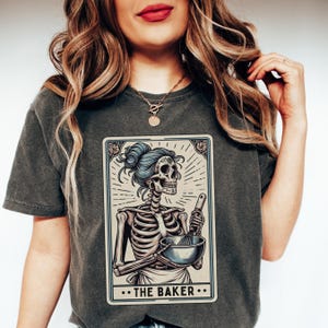 May include: A dark gray t-shirt with a graphic of a skeleton holding a bowl and whisk. The graphic is in the style of a tarot card and reads "...THE BAKER..."