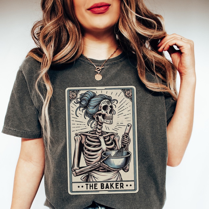 May include: A dark gray t-shirt with a graphic of a skeleton holding a bowl and whisk. The graphic is in the style of a tarot card and reads "...THE BAKER..."