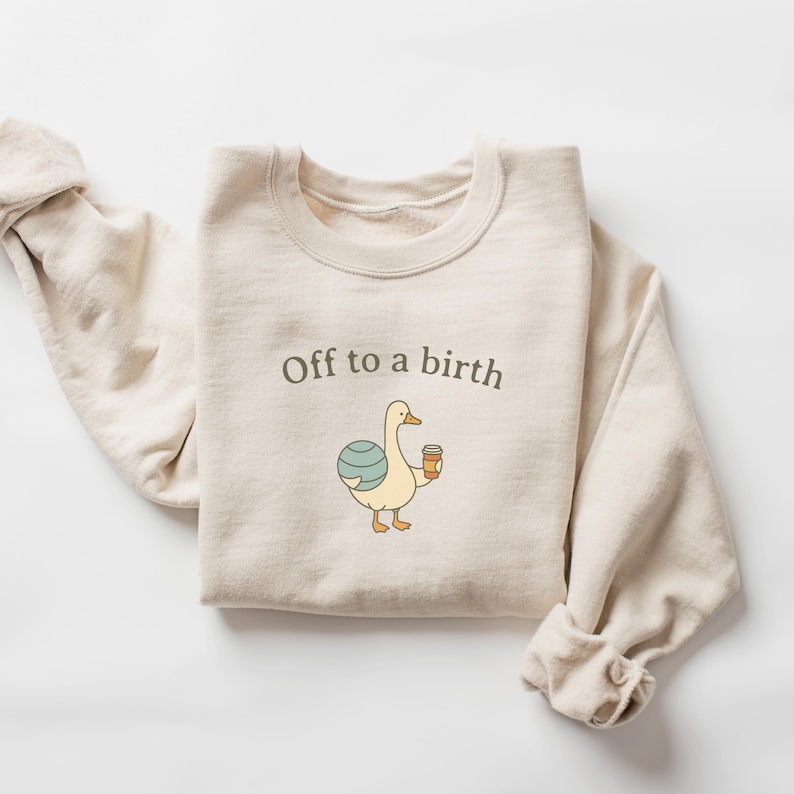 May include: Cream-colored sweatshirt with the text "Off to a birth." The graphic features a cartoon duck holding a coffee cup and a blue and white ball. The sleeves are rolled up.