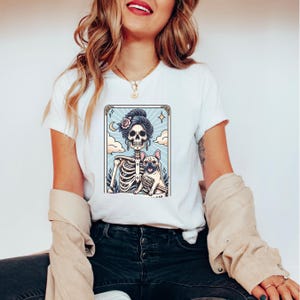 May include: White t-shirt featuring a colorful graphic of a skeleton with a French Bulldog. The skeleton has a floral hairpiece and is set against a light blue background with clouds and stars. The design is framed with a gold border.