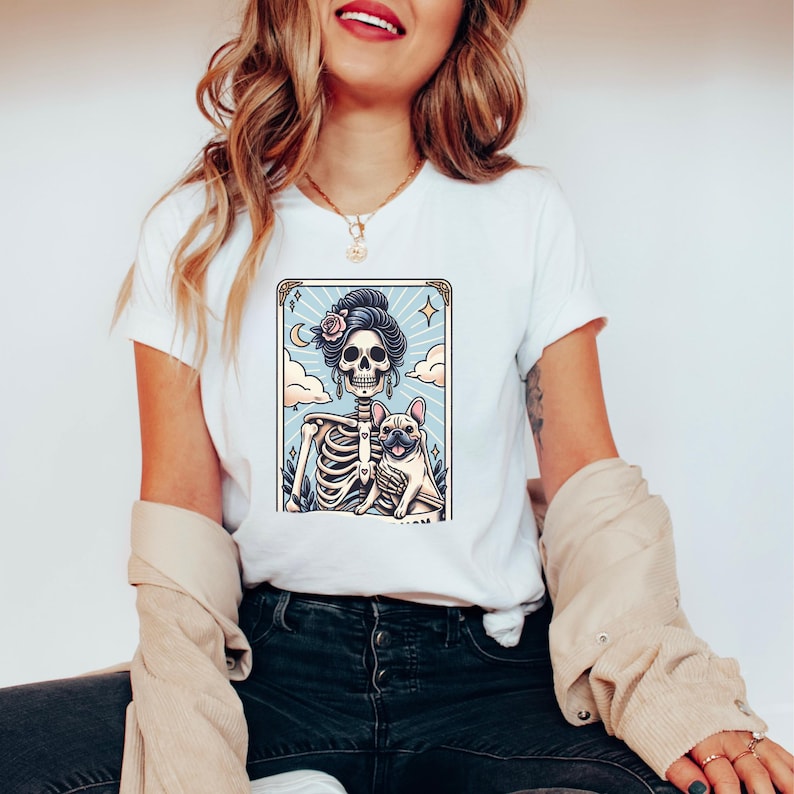May include: White t-shirt featuring a colorful graphic of a skeleton with a French Bulldog. The skeleton has a floral hairpiece and is set against a light blue background with clouds and stars. The design is framed with a gold border.