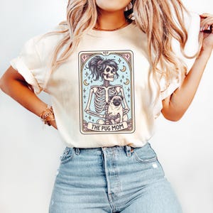 Puede incluir: Una camiseta blanca con un gr&aacute;fico de un esqueleto sosteniendo un pug. El gr&aacute;fico est&aacute; en estilo de carta de tarot con el texto "The Pug Mom" en la parte inferior.