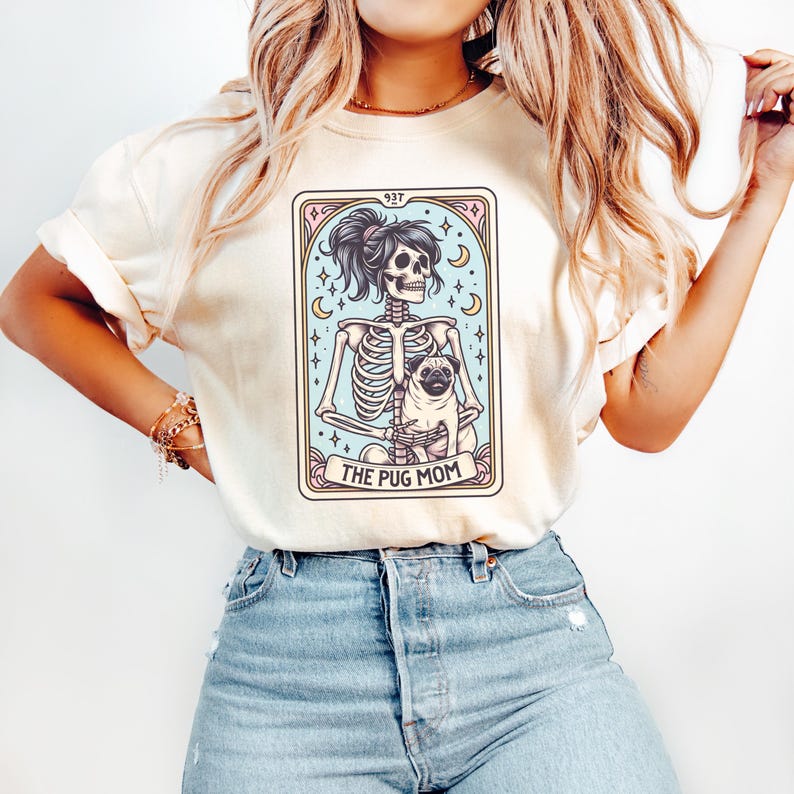 Puede incluir: Una camiseta blanca con un gr&aacute;fico de un esqueleto sosteniendo un pug. El gr&aacute;fico est&aacute; en estilo de carta de tarot con el texto "The Pug Mom" en la parte inferior.