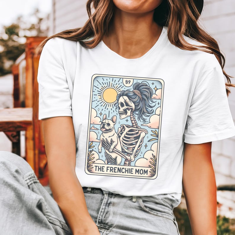 May include: White t-shirt with a graphic of a skeleton holding a French bulldog. The graphic is in a tarot card style with the text "The Frenchie Mom" at the bottom.