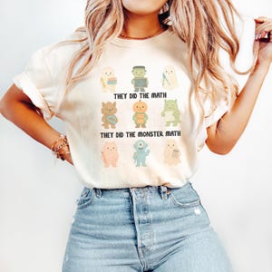 May include: Cream-colored t-shirt featuring a colorful graphic of cartoon monsters. The shirt has the text "THEY DID THE MATH" and "THEY DID THE MONSTER MATH". The monsters are in various colors and shapes.