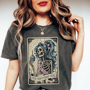 May include: A grey t-shirt featuring a graphic of a skeleton holding a cupcake with a lit candle. The skeleton is wearing a pink sash that says "It's my 30th" and the graphic is surrounded by a gold frame with the text "The Birthday Girl".