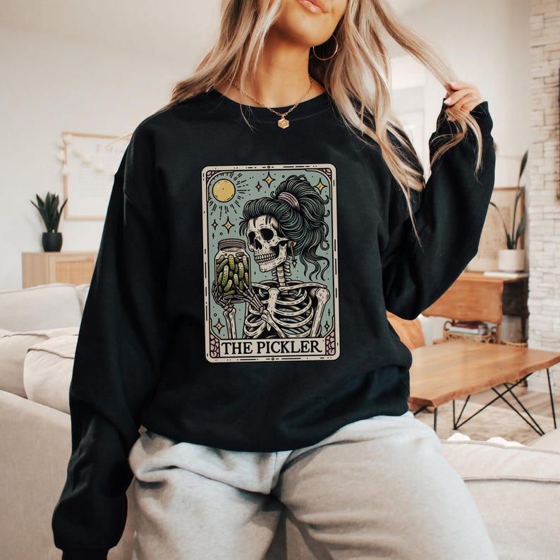May include: Black sweatshirt featuring a tarot card-style graphic. The graphic depicts a skeleton holding a jar of pickles, with the text "THE PICKLER" at the bottom. The design includes a moon, stars, and a decorative border.