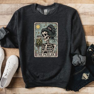 May include: Dark gray sweatshirt featuring a tarot card-style graphic. The graphic depicts a skeleton holding a jar of pickles, with the text "THE PICKLER" at the bottom. The skeleton has teal hair and is set against a light blue background.