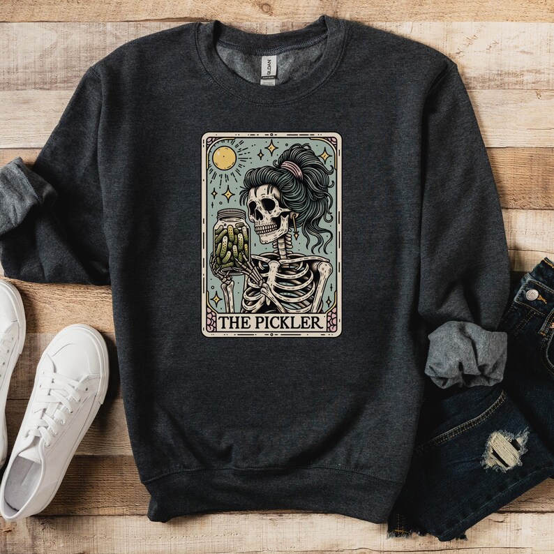 May include: Dark gray sweatshirt featuring a tarot card-style graphic. The graphic depicts a skeleton holding a jar of pickles, with the text "THE PICKLER" at the bottom. The skeleton has teal hair and is set against a light blue background.