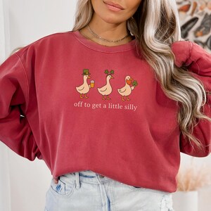 May include: A maroon sweatshirt with a whimsical design featuring three cartoon ducks. Each duck is adorned with St. Patrick's Day themed accessories and holding a beverage or shamrocks. The text "off to get a little silly" is printed below the ducks.