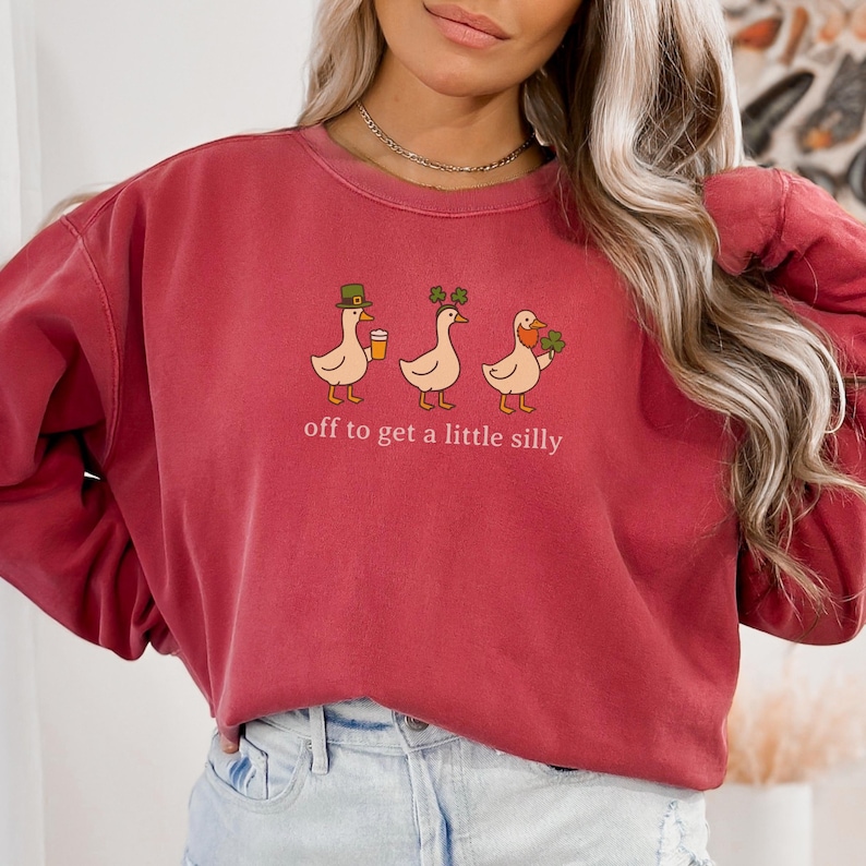 May include: A maroon sweatshirt with a whimsical design featuring three cartoon ducks. Each duck is adorned with St. Patrick's Day themed accessories and holding a beverage or shamrocks. The text "off to get a little silly" is printed below the ducks.