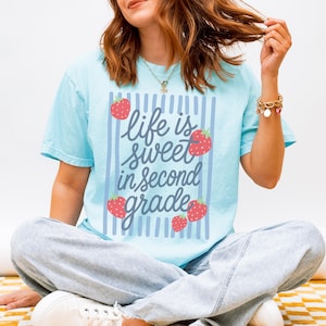 K&ouml;nnte beinhalten: Hellblaues T-Shirt mit dem Text "Life is sweet in second grade" in verspielter Schrift, umrahmt von vertikalen blauen Streifen und roten Erdbeer-Illustrationen. Das Shirt ist ein l&auml;ssiger Stil mit kurzen &Auml;rmeln.
