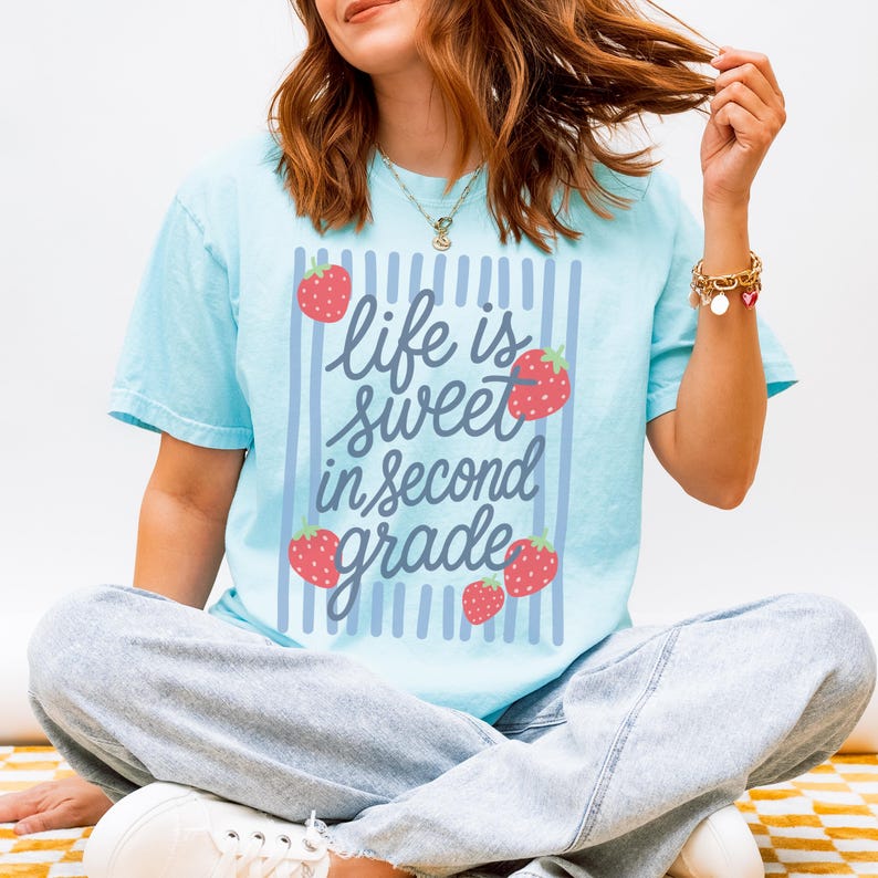 K&ouml;nnte beinhalten: Hellblaues T-Shirt mit dem Text "Life is sweet in second grade" in verspielter Schrift, umrahmt von vertikalen blauen Streifen und roten Erdbeer-Illustrationen. Das Shirt ist ein l&auml;ssiger Stil mit kurzen &Auml;rmeln.