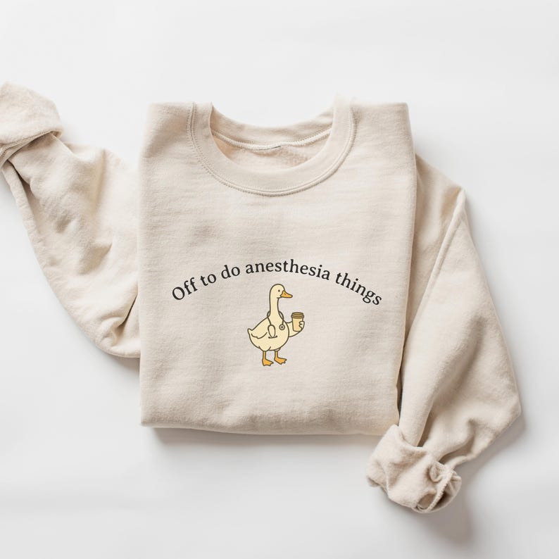 Puede incluir: Sudadera color crema con la frase "Off to do anesthesia things" impresa en negro. Un pato de dibujos animados con estetoscopio y taza de caf&eacute; est&aacute; centrado debajo del texto. La sudadera tiene cuello redondo y mangas largas.