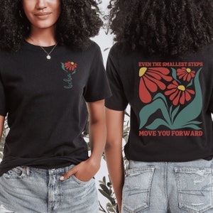 May include: Two black t-shirts with a floral design and text. The front shirt has a single flower with the word "bebo" written below it. The back shirt has a larger floral design with the text "Even the smallest steps move you forward".