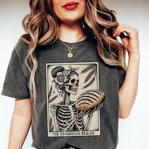 May include: A dark gray t-shirt with a graphic of a skeleton holding a loaf of bread. The graphic is in a vintage style and includes the text "The Sourdough Dealer".