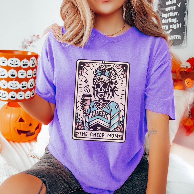 Pu&ograve; includere: Maglietta viola con una grafica di uno scheletro che indossa un costume da cheerleader e tiene una tazza di caff&egrave;. La grafica &egrave; in stile carta dei tarocchi con il testo "The Cheer Mom" in basso.