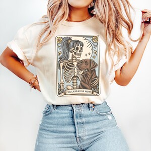 May include: A white t-shirt with a graphic of a skeleton holding a labrador retriever. The graphic is in a tarot card style with the text "THE LABRADOR MOM" at the bottom.