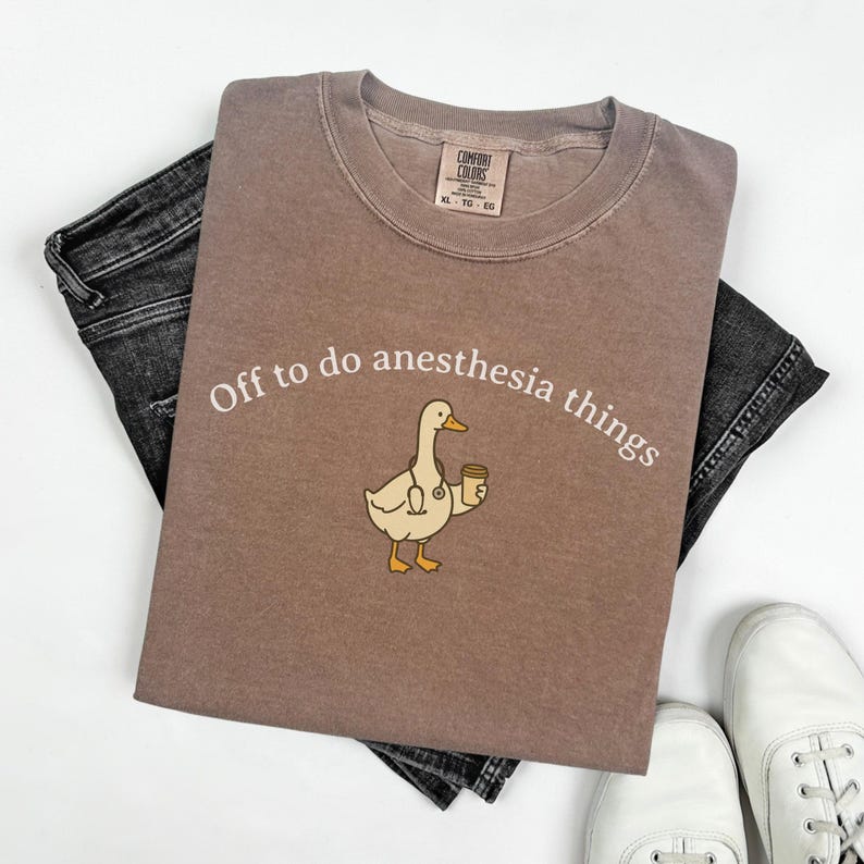 May include: A brown Comfort Colors t-shirt with the text "Off to do anesthesia things". The shirt features a cartoon duck holding a coffee cup and stethoscope. The shirt is paired with black jeans and white shoes.