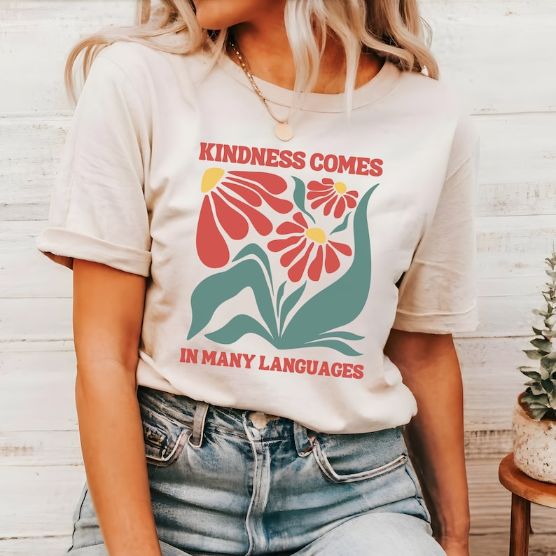 May include: A cream colored t-shirt with a graphic of red flowers with yellow centers and green leaves. The text "KINDNESS COMES IN MANY LANGUAGES" is printed in red on the shirt.