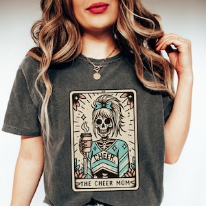 Pu&ograve; includere: Una maglietta grigio scuro con una grafica di uno scheletro che indossa un'uniforme da cheerleader e tiene una tazza di caff&egrave;. La grafica &egrave; in stile vintage e include il testo "Cheer" e "The Cheer Mom".