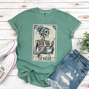 May include: A green t-shirt with a vintage-style illustration of a skeleton holding a bowl and whisk. The illustration is in a card-like frame with the text "The Baker" below it.